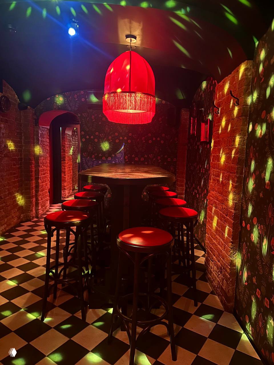 Высокий круглый стол и ряд табуретов в узком коридоре с кирпичными стенами, красной лампой и множеством зелёных световых пятен — интерьерный проход для больших компаний.