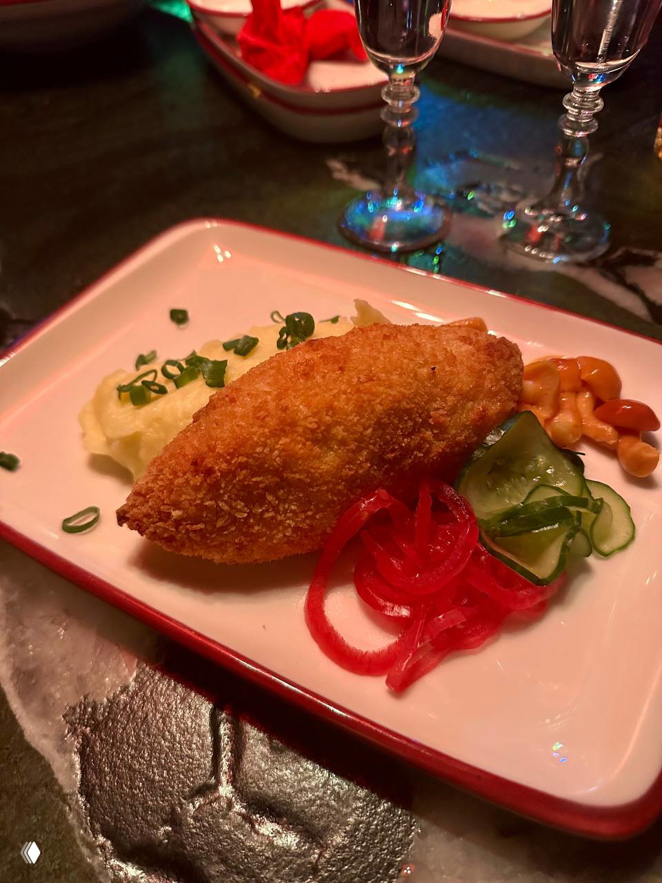 Жареная закуска в панировке на белой прямоугольной тарелке с гарниром и соленьями, типичная закуска рюмочной кухни, снятая крупным планом при тёплом освещении.