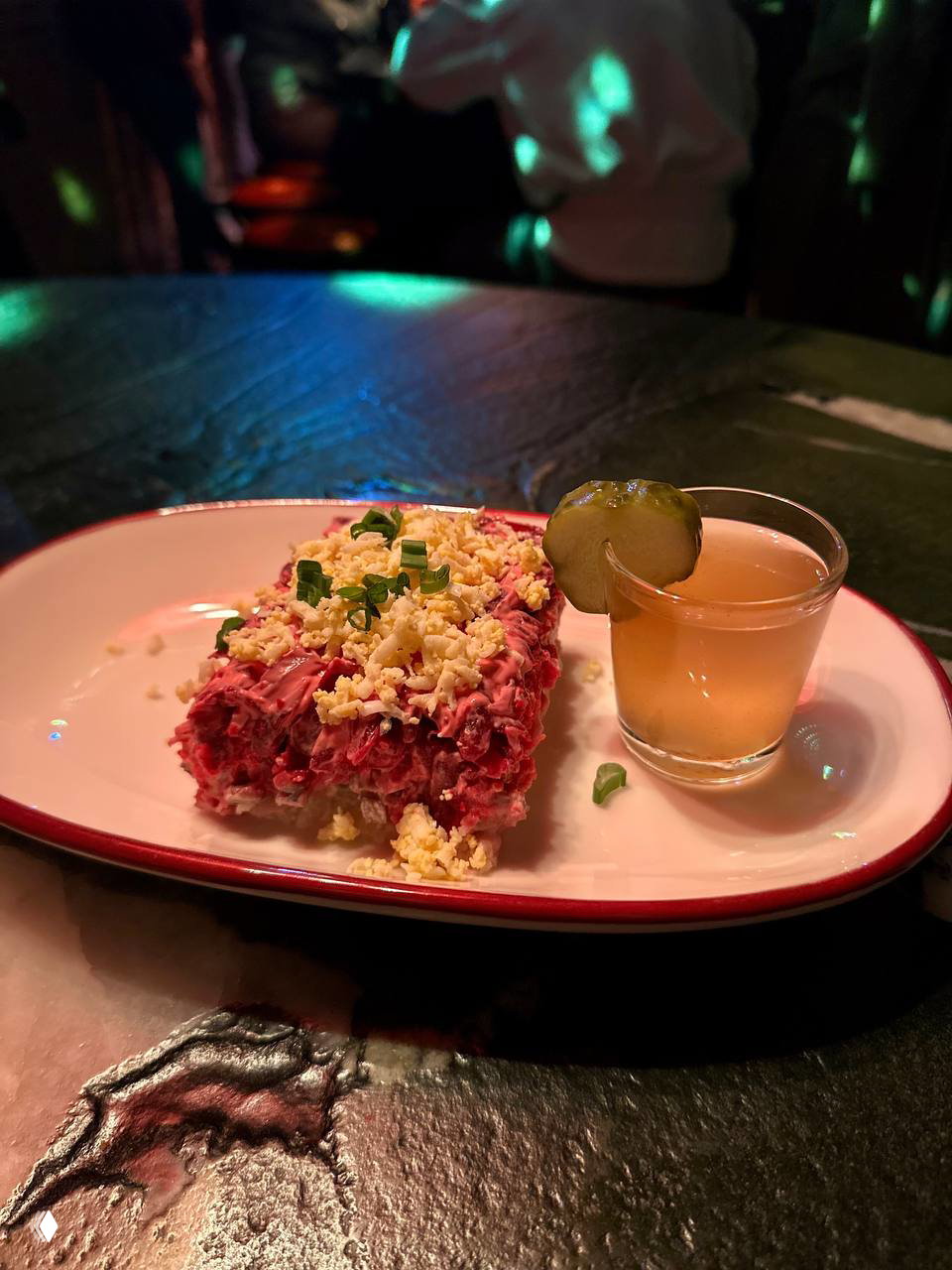 Порция салата в ретро‑подавке и маленькая рюмка с закуской на тёмном мраморном столе: крупный план еды на фоне красных огней и диско‑бликов интерьера.