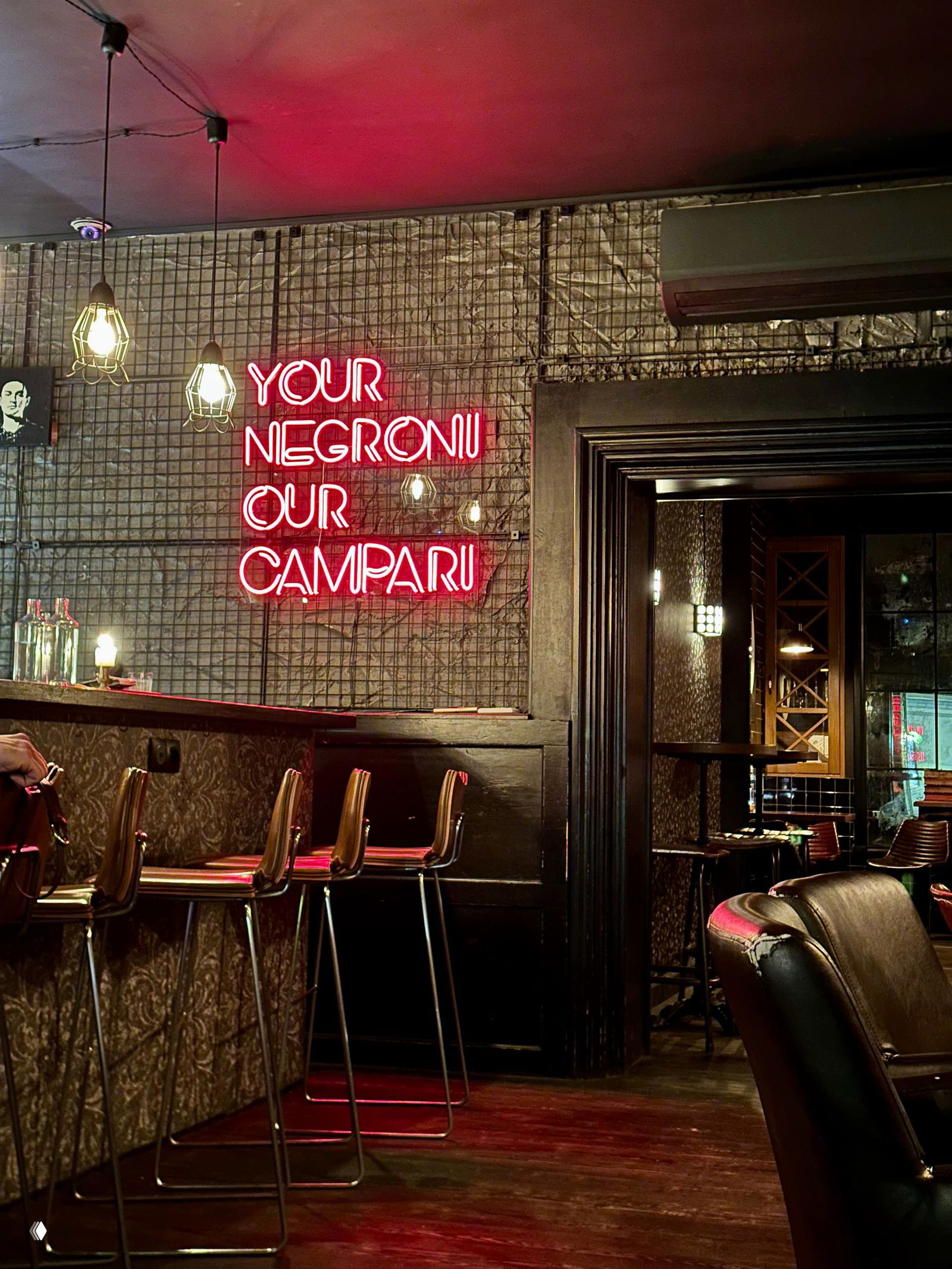 Барная стойка и неоновая вывеска «YOUR NEGRONI OUR CAMPARI» в тёплом красно-янтарном свете, видно барные стулья и часть интерьера Imbibe.