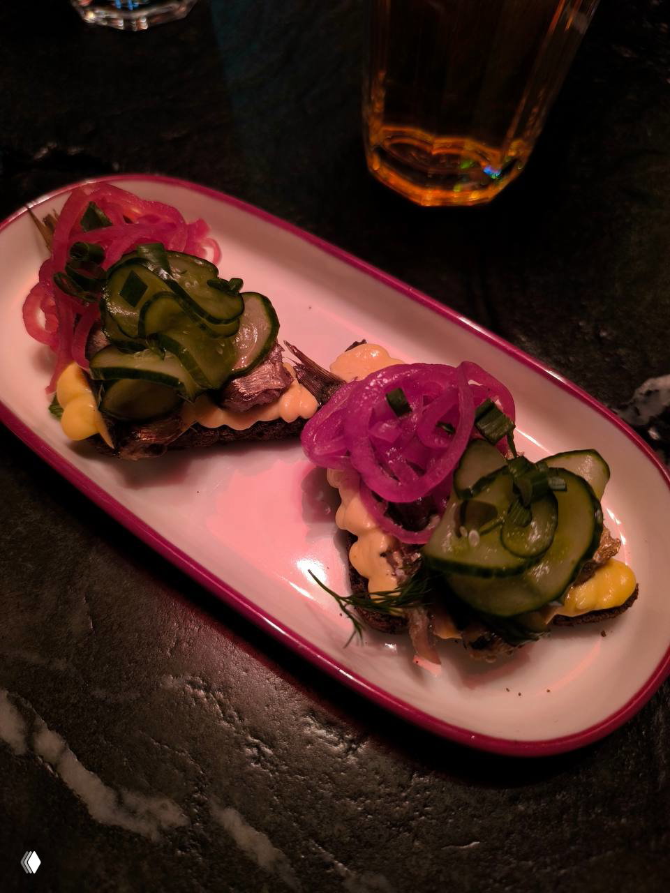 Две небольшие закусочные тарелки с маринованными огурцами, луком и кусочками хлеба под тёплым красным освещением — закуски к рюмке в типичной подаче заведения.