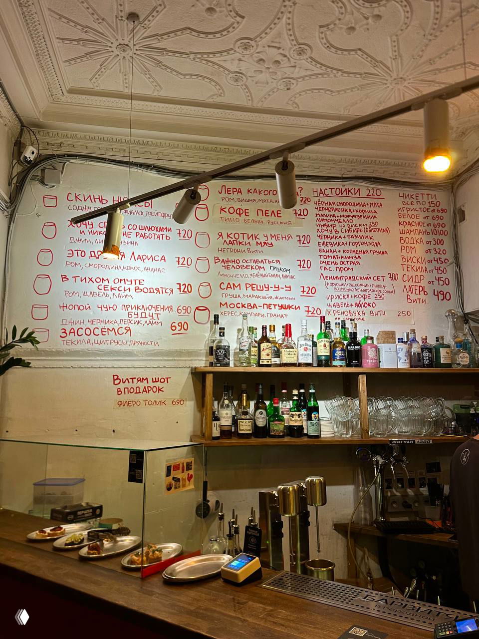 Барная стойка и стена с рукописным меню в баре «Витя»: бутылки, подносы с закусками и светильники в уютной непринуждённой атмосфере.