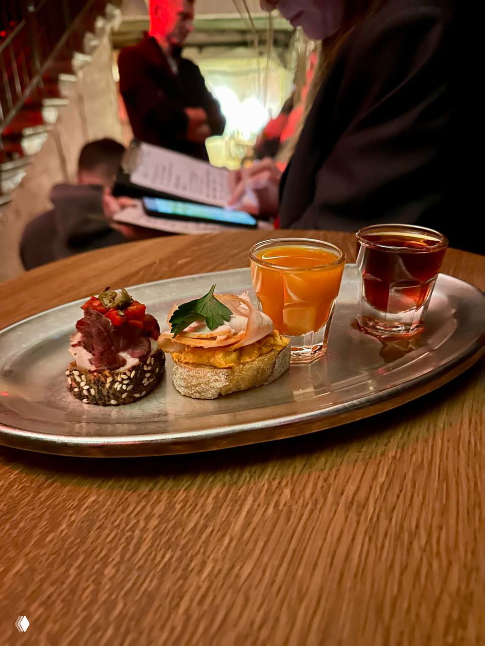 Металлический поднос с двумя рюмками и мини-закусками на деревянном столе в баре «Витя», на заднем плане посетители и лестничный пролет.