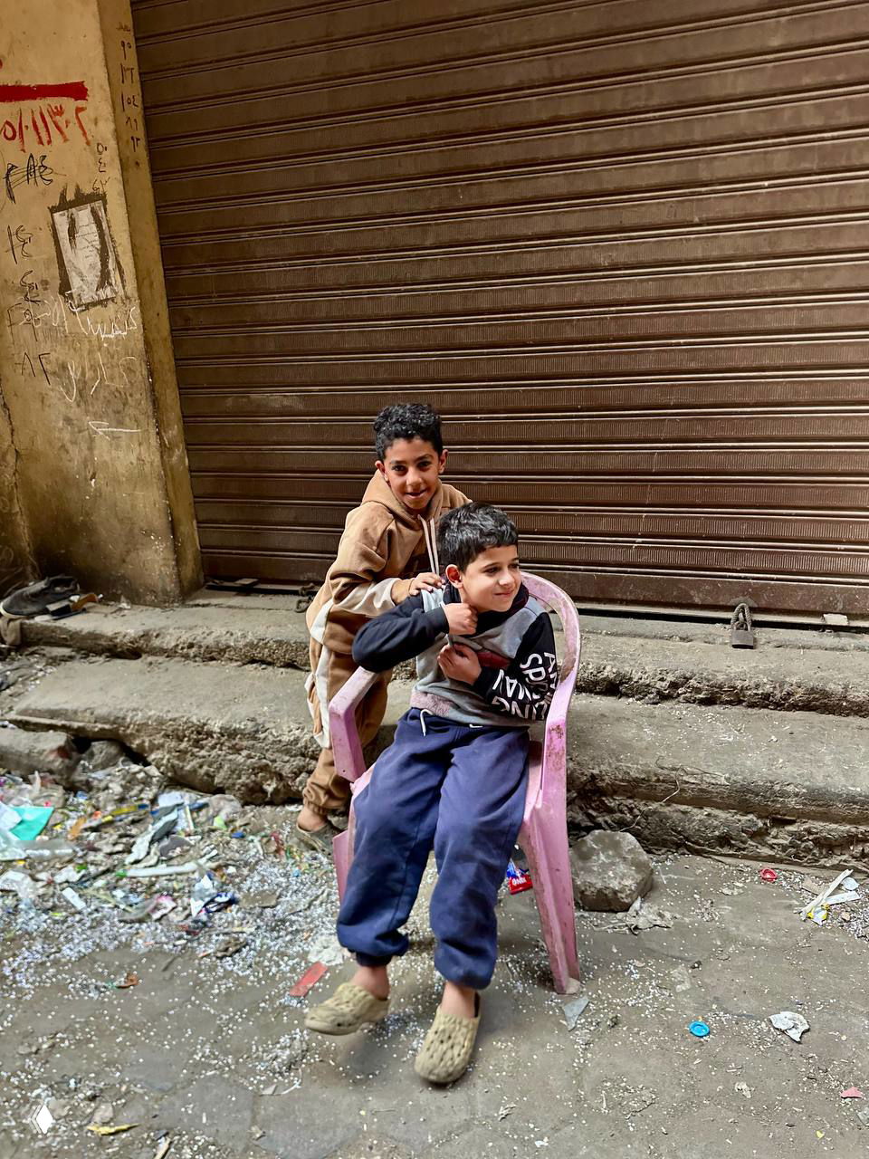 Двое играющих мальчиков в грязном переулке на фоне мусора и закрытого металлического роллетного входа — момент повседневного уличного быта.