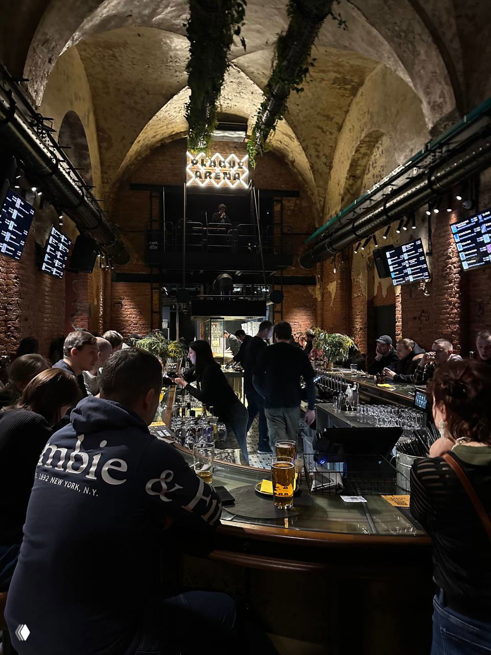 Посетители за барной стойкой Plague Arena: люди пьют пиво и общаются при низком освещении под неоновым логотипом и сводчатым потолком.