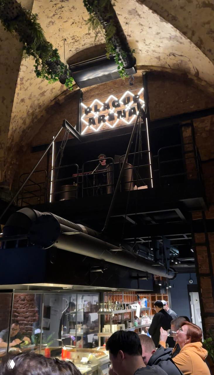 Неоновый логотип Plague Arena на мезонине над барной зоной, сцена и техническое оборудование видны на фоне арочных потолков и растений.