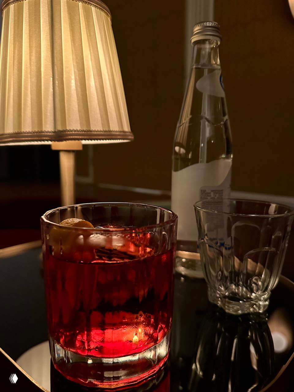 Коктейль в низком стакане на зеркальном столике кинотеатра Michele: красный напиток, рядом бутылка воды и пустой бокал, тёплая лампа создаёт уют.
