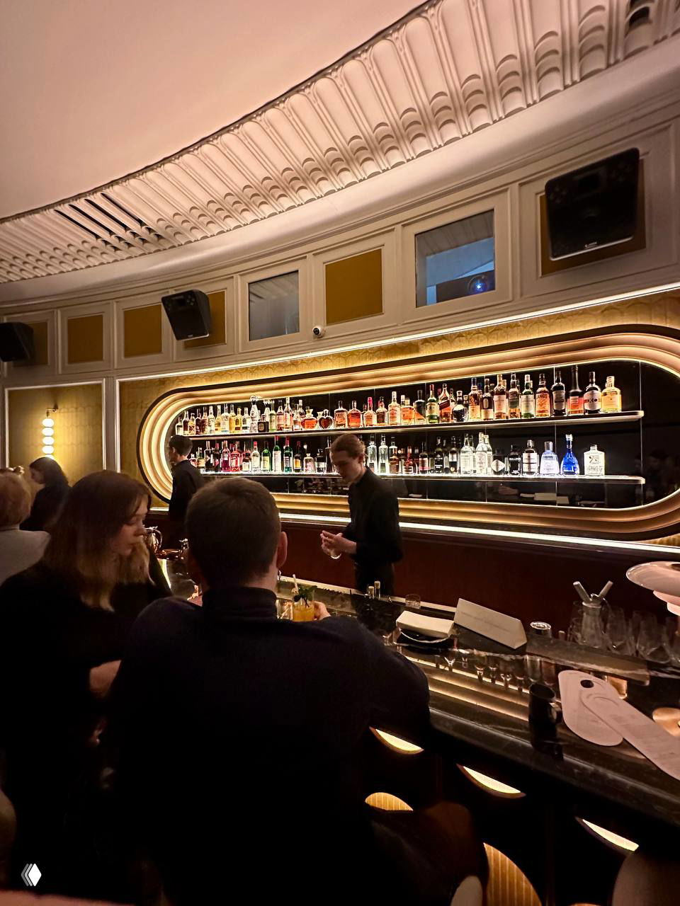 Барная стойка в Michele: подсвеченная полка с ассортиментом бутылок, бармены за работой и посетители у стойки в тёплой атмосферной подсветке.