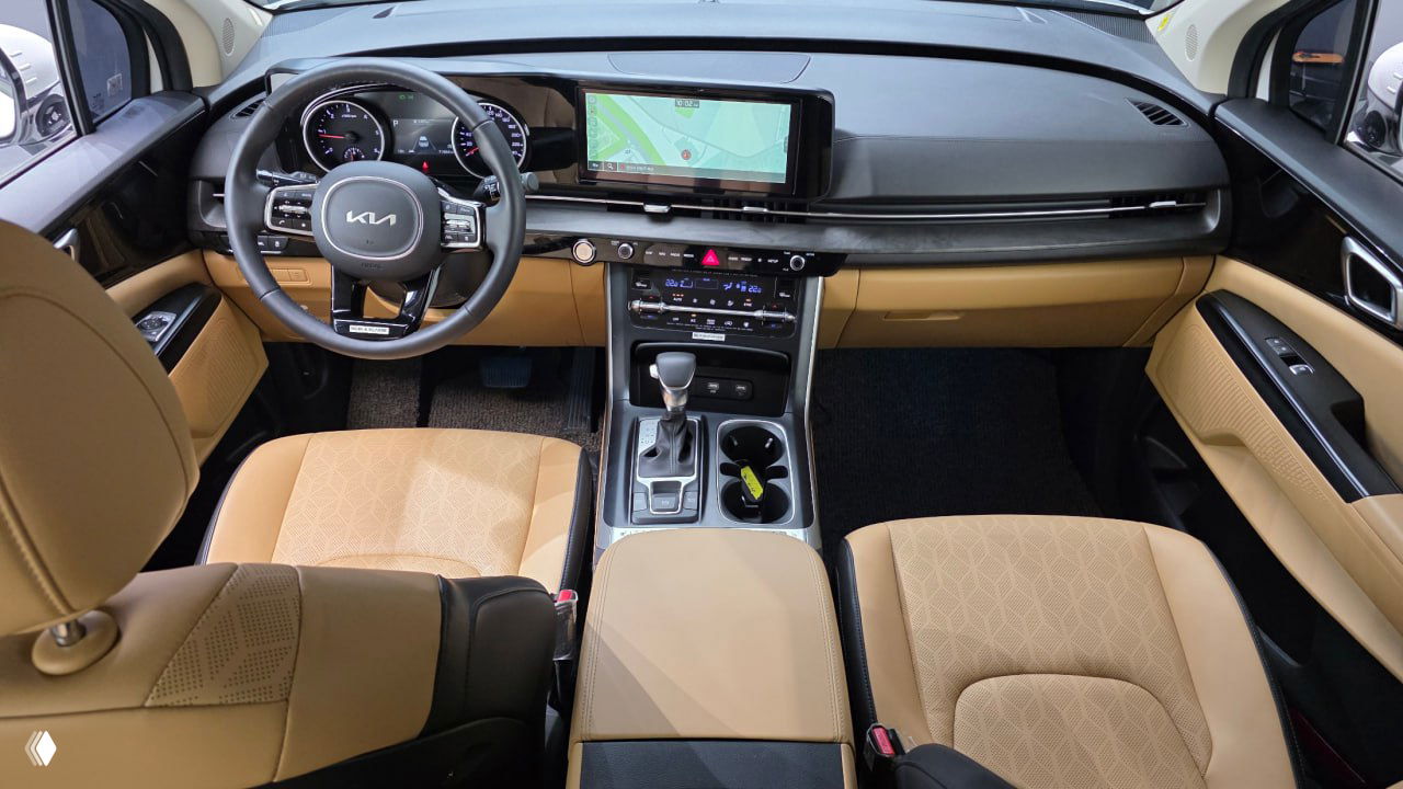 Интерьер Kia Carnival 2023: торпедо с большим экраном навигации, руль с логотипом KIA и бежевые передние кожаные сиденья крупным планом.