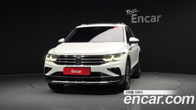 Белый Volkswagen Tiguan (Prestige) 2021 — вид спереди в студийном освещении, включённые фары и фирменный логотип Encar на фоне чёрной стены.