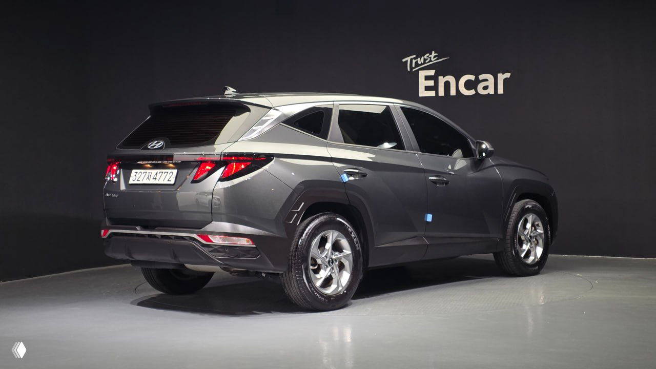 Задний трёхчетвертной вид Hyundai Tucson NX4 в сером цвете на фоне логотипа Encar, видна задняя часть, фары и колёса автомобиля.