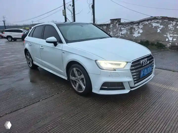 Белая Audi A3/Q2L 2021 года на мокрой парковке в городе, вид под углом с включёнными фарами, демонстрация дисков и бамперов