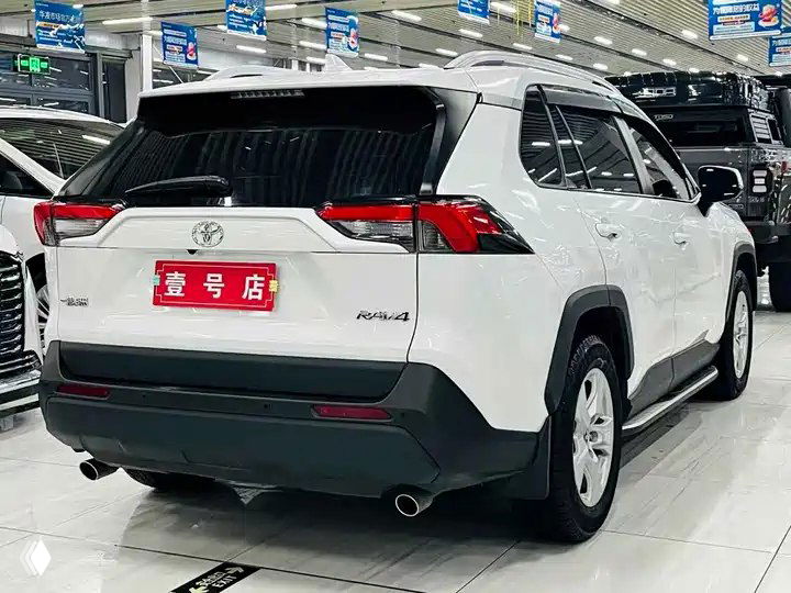Белый Toyota RAV4 Urban Edition 2023 в автосалоне — вид сзади и сбоку, отчётливо видны фонари, багажник, задний бампер и выхлопная труба.