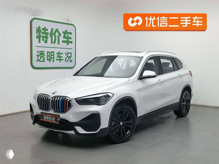 Белый BMW X1 2021 года в выставочном зале с ценниками и логотипами площадки на заднем плане, ракурс 3/4 спереди
