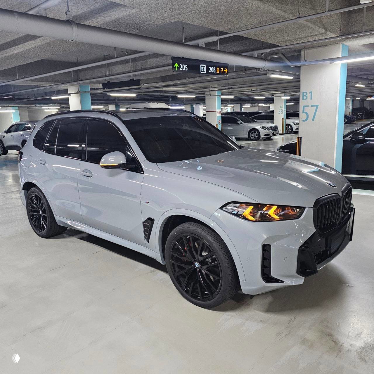 Серый BMW X5 xDrive40d 2026 года на подземной парковке в Южной Корее, вид спереди с включёнными фарами и чёрными дисками.