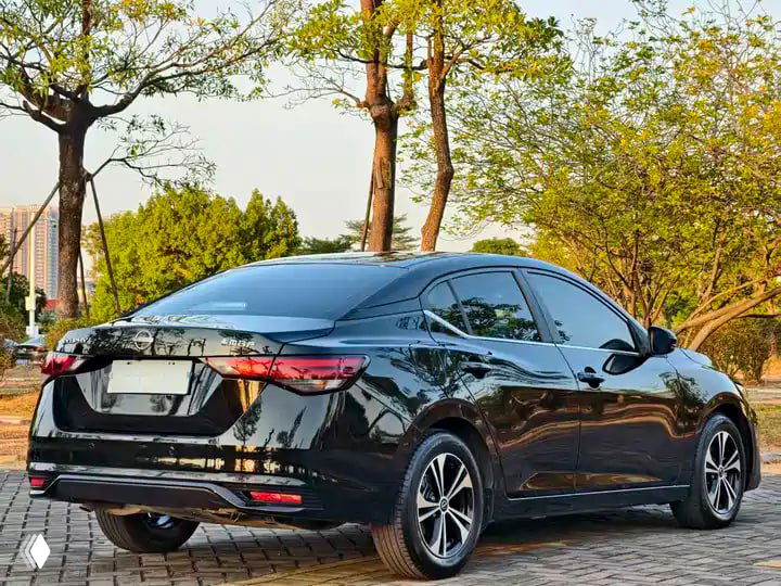 Задняя часть чёрного NISSAN Sylphy (рестайлинг) 2023, вид сзади под углом, видна форма багажника и фон с деревьями и городской инфраструктурой.