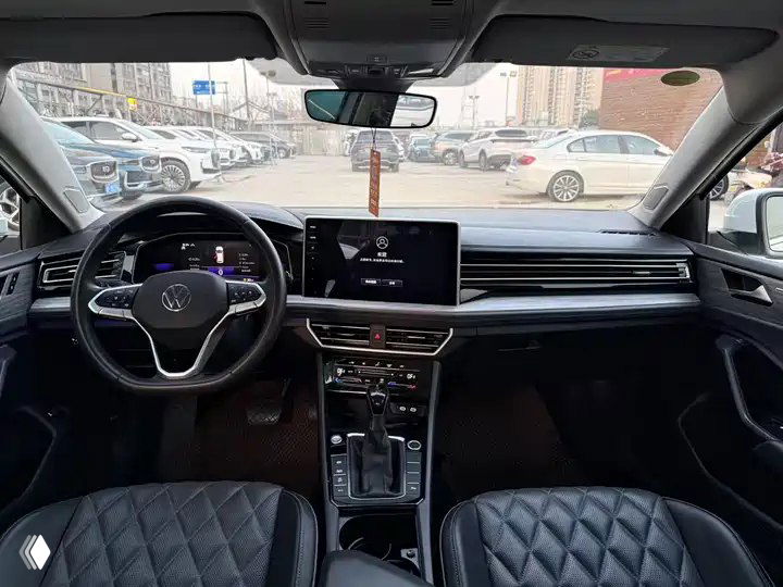 Интерьер Volkswagen Sagitar: руль, панель приборов и мультимедиа-экран; видны передние сиденья с ромбовой прострочкой и стоянка за лобовым.