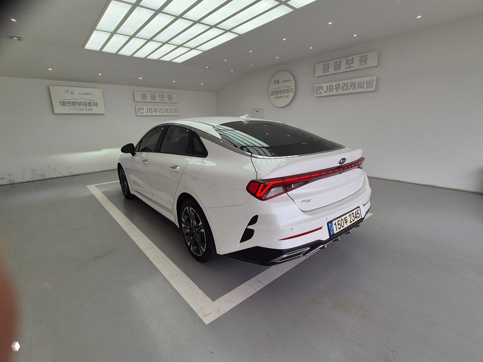 Белый KIA K5 2021 Noblesse, вид сзади в выставочном зале; интегрированные фонари, задний бампер и корейский регистрационный номер видны.