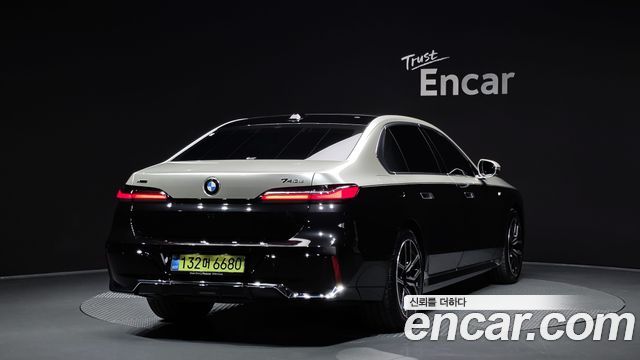 Вид сзади BMW 7 Series 740d xDrive M Sport 2025 чёрного цвета на поворотной площадке шоурума с подсветкой и логотипом Encar на стене.
