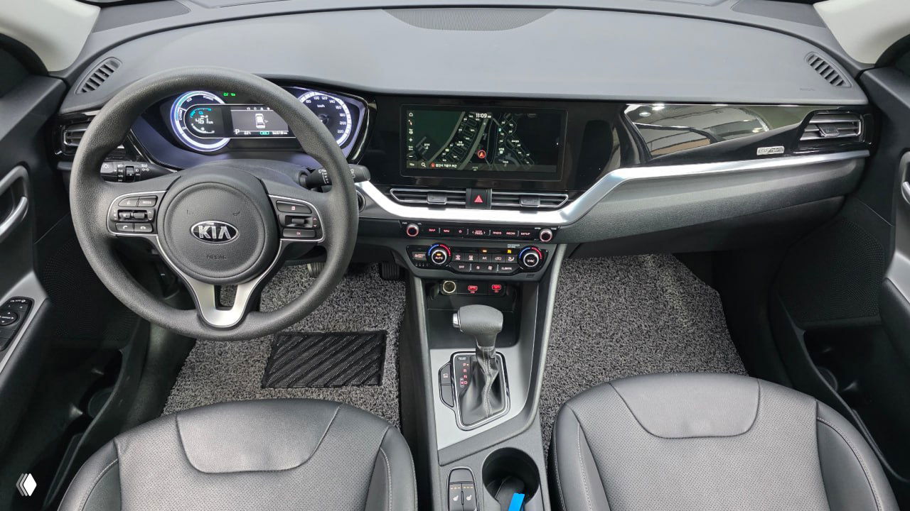 Салон KIA The New Niro — руль с эмблемой KIA, приборная панель и центральный экран, вид на передние сиденья и консоль.