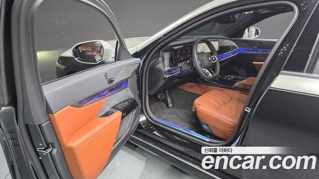 Интерьер BMW 7 Series 740d xDrive M Sport 2025: открытая передняя дверь, коричневые кожаные сиденья и панель приборов с подсветкой в шоурумной обстановке.