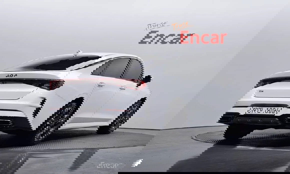 Задний три четверти ракурс белого KIA K5 2022 с видимой южнокорейской номерной пластиной и логотипом Encar на фоне студийной стены.