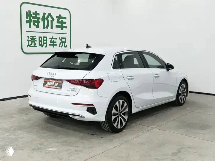 Задняя часть белого Audi A3 Sportback 2022 в шоуруме, виден багажник, задние фонари и рекламные стенды на стене позади автомобиля