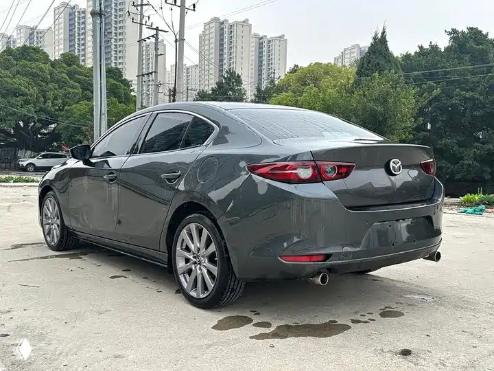 Задний трёхчетвертной ракурс серой Mazda3 Axela 2022 на фоне многоэтажек; видны фонари, бампер и выхлопная труба.