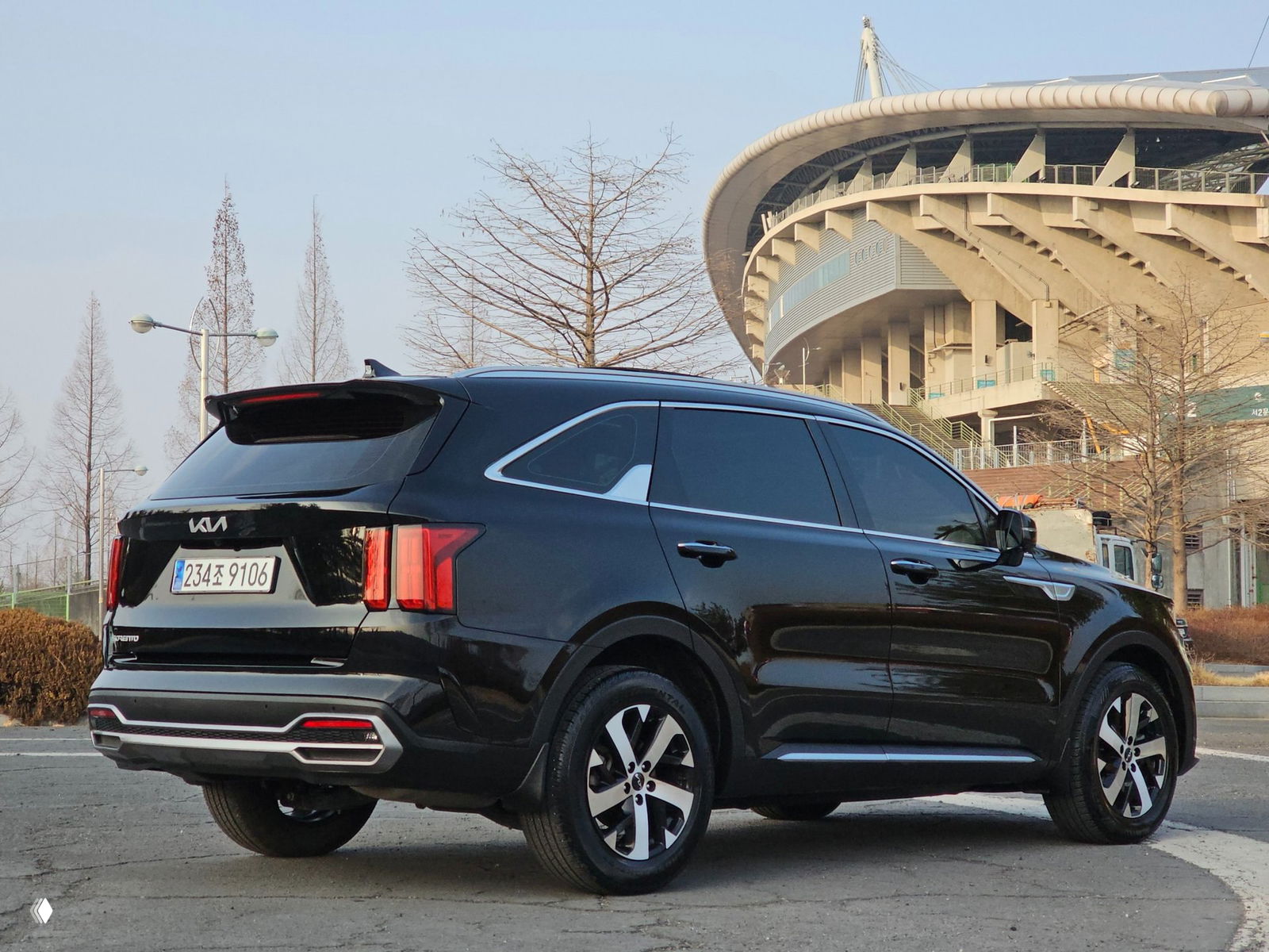 Задний три четверти ракурс чёрного KIA Sorento 2023 на фоне стадиона, видно форму кормы, фонари и бампер автомобиля.
