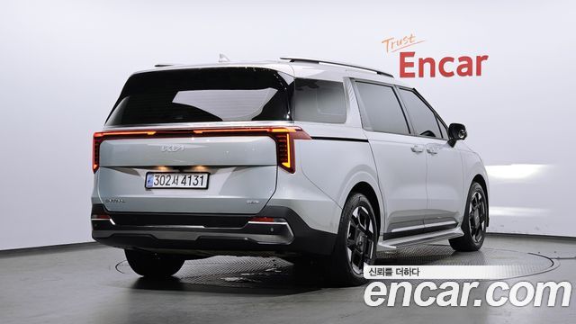 Вид сзади Kia Carnival HEV 2024 в студийном павильоне Encar, видно горизонтальную светодиодную подсветку и заднюю часть машины.