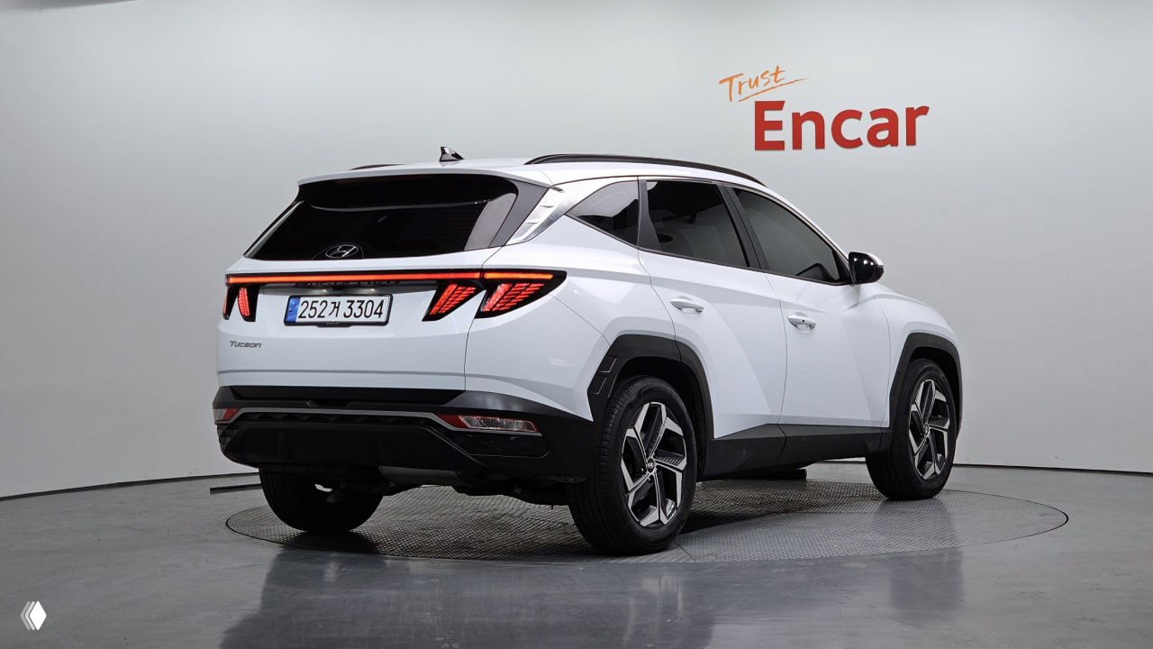 Фотография белого Hyundai Tucson 2023 года в шоуруме Encar, вид сзади и боковой части, видна кормовая оптика, номерная табличка и логотип стенда.