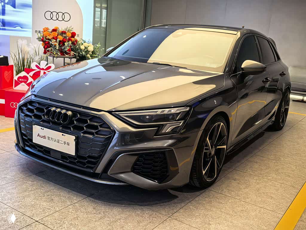 Уголовой вид Audi A3 Sportback 2022 серого цвета на стоянке шоурума, заметны колёсные диски и спортивные линии корпуса автомобиля.