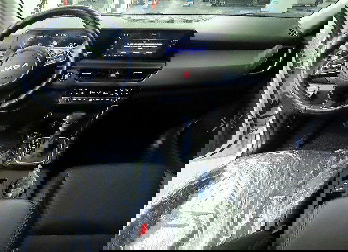 Интерьер KIA Seltos 2023: салон с рулём и мультимедиа дисплеем, видна центральная консоль, рычаг КПП и защитные плёнки на сиденьях.