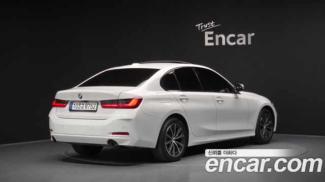 Белый BMW 3 Series 320i 2024 года в салоне выставочного пространства Encar — вид сзади и боковой профиль автомобиля.