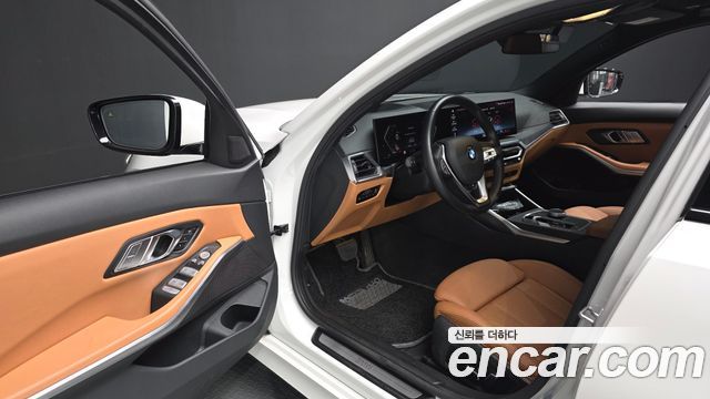 Интерьер BMW 3 Series 320i 2024: открытая передняя дверь, коричневая отделка сидений и приборная панель, вид водительского места.