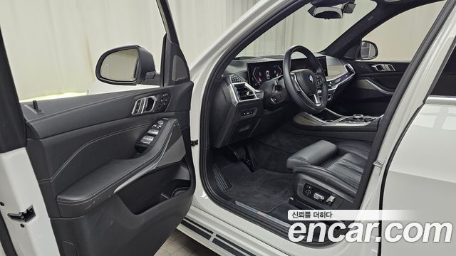Интерьер BMW X7 2024: открытая дверь, водительское сиденье, приборная панель и руль, чёрная отделка, обзор на центральную консоль