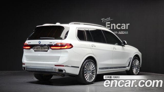 Белый BMW X7 xDrive 40i 2024 в шоуруме, вид сзади-слегка-сбоку, видны фонари и багажник, водяной знак и студийное освещение