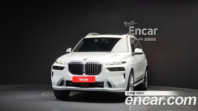 Белый BMW X7 xDrive 40i 2024 в шоуруме, крупный план передней части, выделены фары и радиаторная решётка, логотип Encar виден на фото
