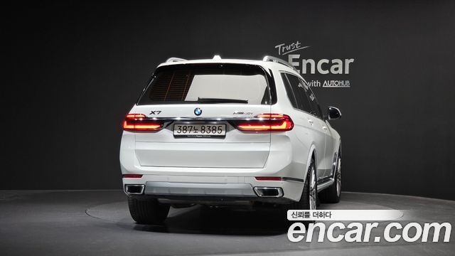 Белый BMW X7 xDrive 40i 2024 вид сзади в шоуруме, подчёркнуты задние фонари и бампер, на фото студийный фон и логотип Encar