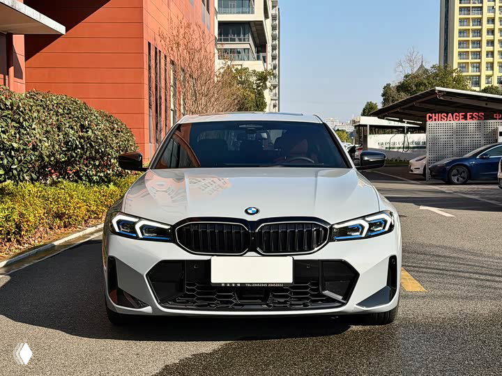 Фронтальный крупный план BMW 3 330Li M 2025: решётка, фары и бампер на фоне городской застройки и парковки в Китае, дневной свет.