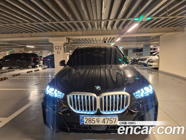 Фронтальный крупный план BMW X5 2026 в подземной парковке: крупная решётка радиатора, светодиодные фары, отражения потолочных ламп на капоте.