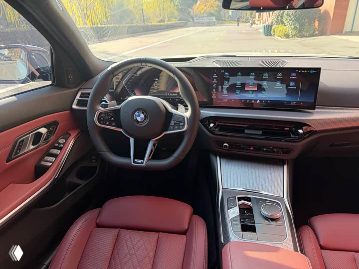 Салон BMW 3 330Li M 2025 — вид с водительского места: мультифункциональный руль, цифровая приборная панель и большой экран мультимедиа, красная обивка.