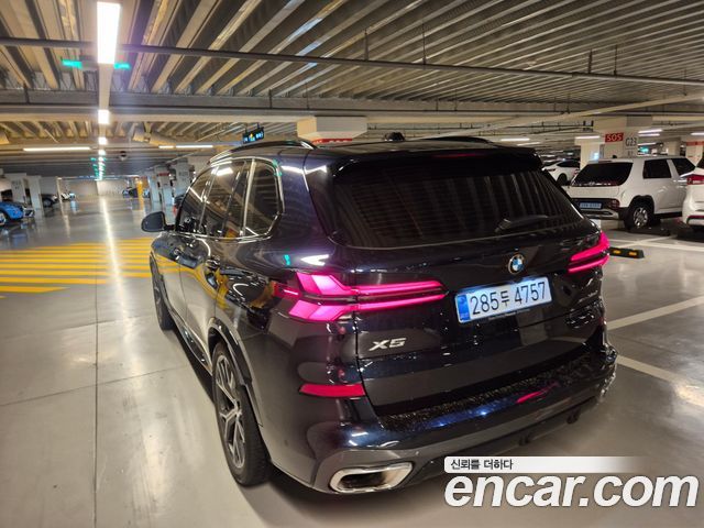 Задний ракурс чёрного BMW X5 xDrive 30d M Sport 2026 в паркинге; видны задние фонари, эмблема X5 и элементы бампера на фоне других авто.