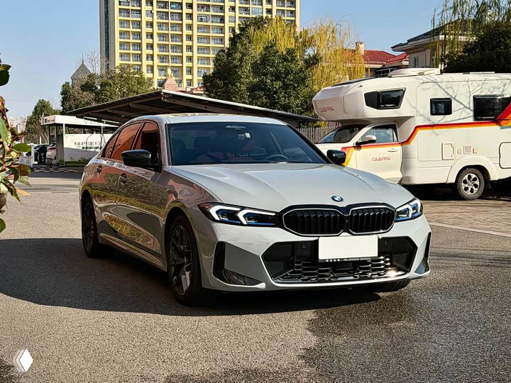 Боковой и передний ракурсы BMW 3 330Li M 2025 на стоянке: виден профиль седана, отражения на кузове и автодом на заднем плане.