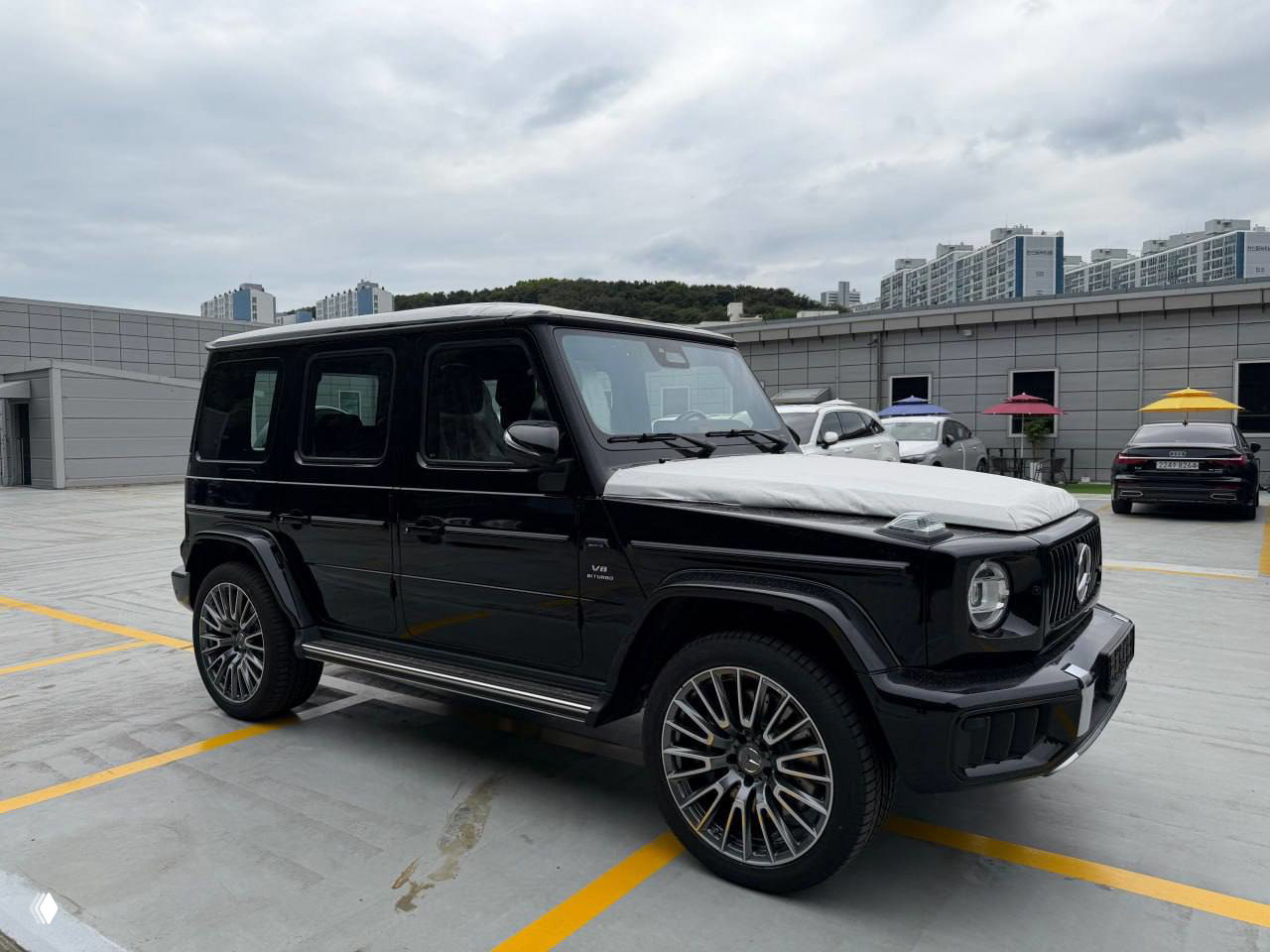 Боковой ракурс чёрного Mercedes‑G63 2026 с крупными легкосплавными дисками, парковочное место на крыше комплекса и жилые дома на заднем плане.