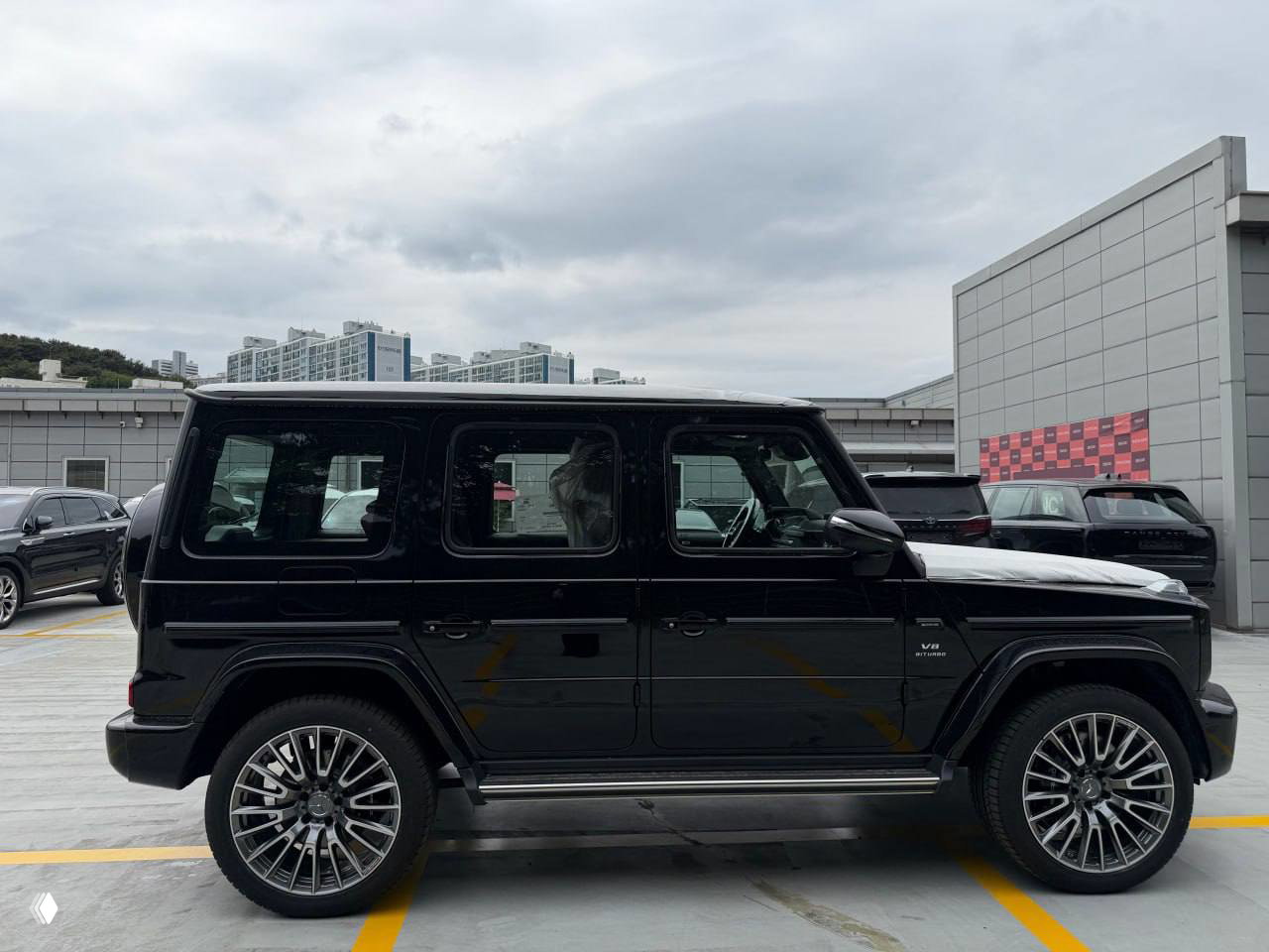 Профильный вид чёрного Mercedes‑AMG G63 2026, хорошо видна эмблема V8 Biturbo на переднем крыле и типичный городской пейзаж позади автомобиля.