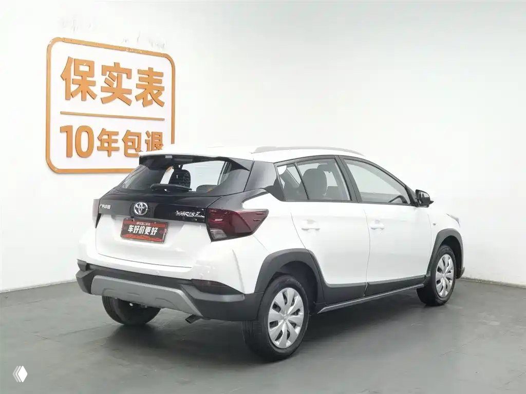 Задний трехчетвертной вид белого Toyota YARiS L 2021 года в шоуруме, видна задняя дверь и бампер, автомобиль на подиуме.