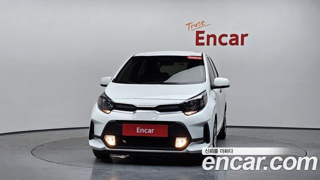 Фронтальный вид белого Kia Morning на студийном фоне; видно переднюю часть кузова, фары и логотип дилера, фото для лота из Кореи.