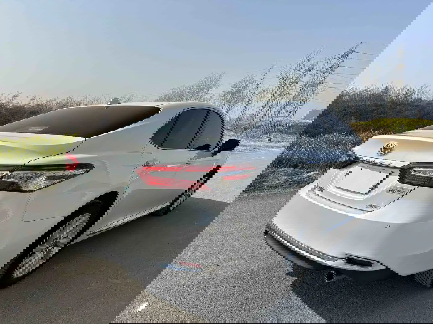 Задняя часть белой Toyota Camry 2023: багажник, задние фонари и правая сторона кузова на дорожном фоне с растительностью и линиями электропередачи.