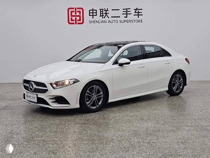 Белый седан Mercedes‑Benz A‑класса снят в шоуруме на нейтральном фоне, вид с левой стороны, чистый пол и стенд дилера.